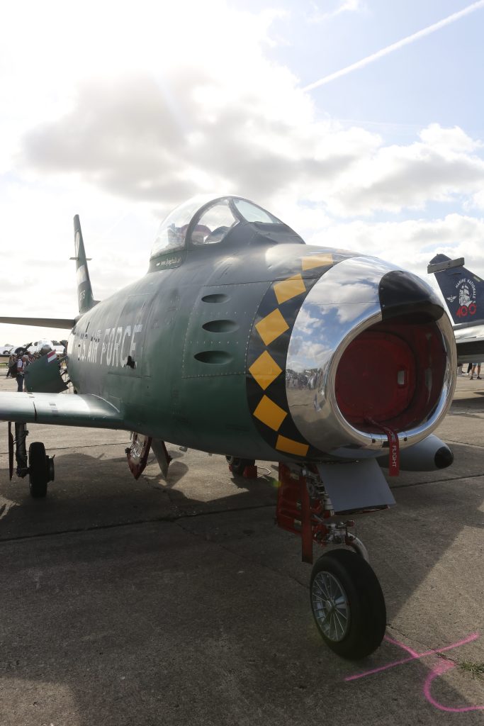 Een militaire jet met een groene en zilveren kleur, uitgerust met een opvallende geel-zwarte standaard. De cockpit is zichtbaar, met wolken en een blauwe lucht op de achtergrond.
