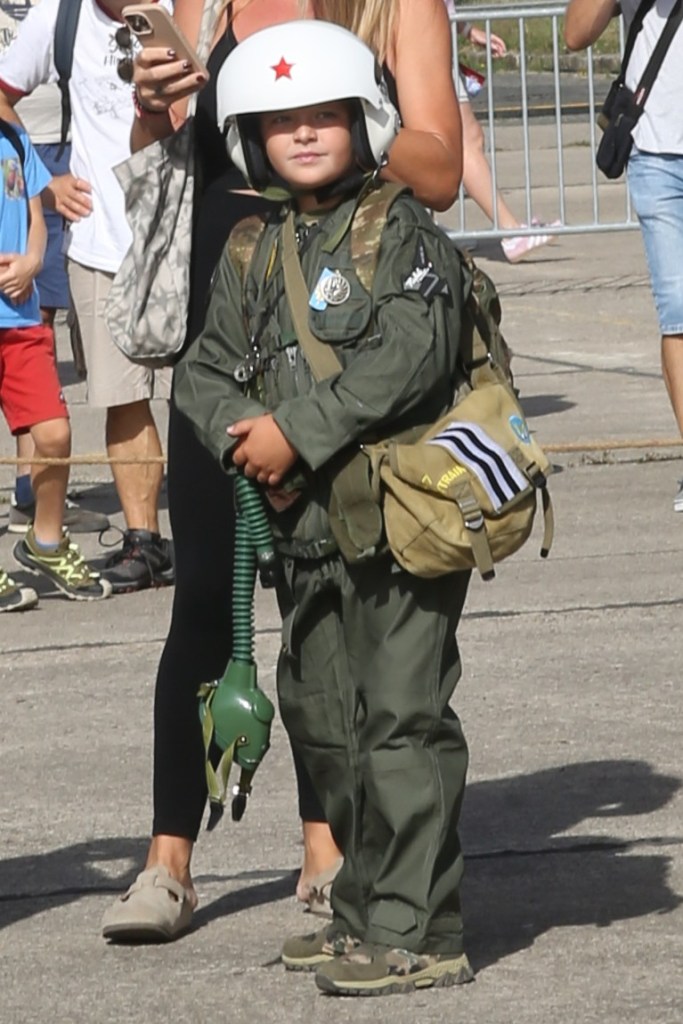 Een jong kind in een militair uniform met een helm en zuurstofmasker, staande naast een volwassene op een druk evenement.