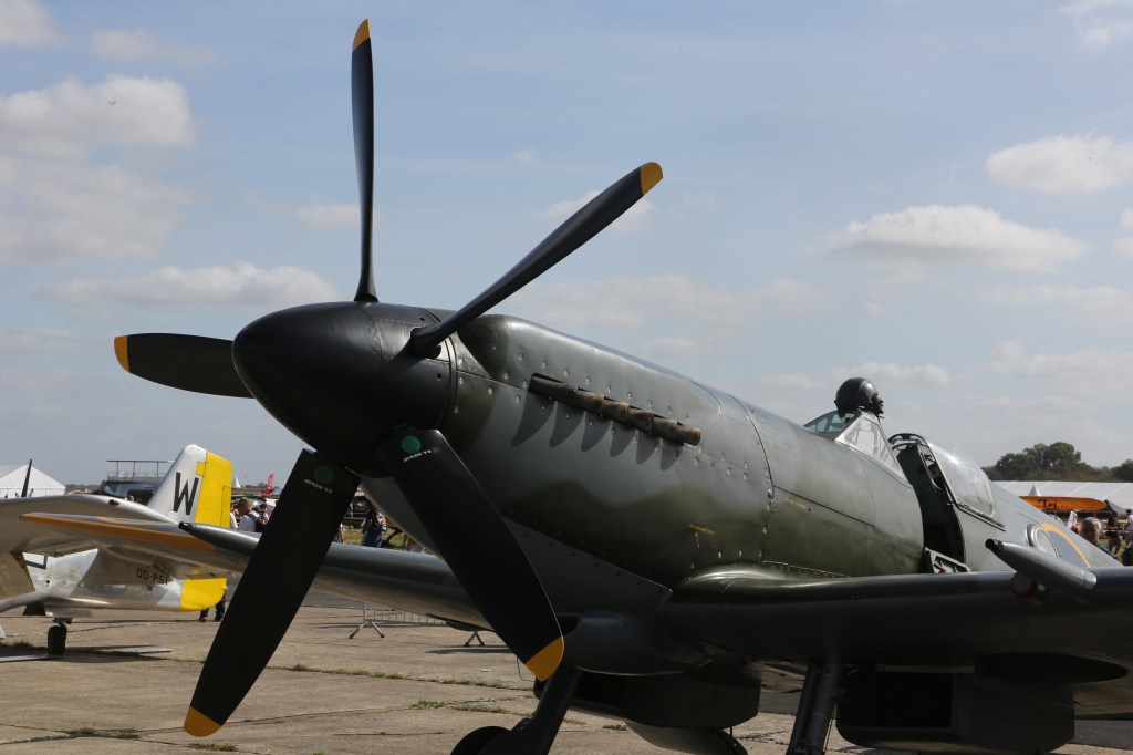Een klassieke jachtvliegtuig met een zwarte propeller en een groene camouflagekleur, weergegeven op een luchtvaartshow met mensen op de achtergrond.