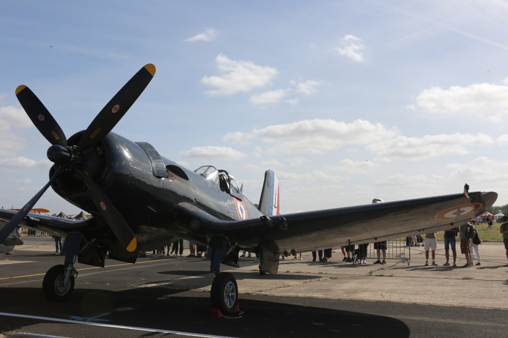 Een historische oorlogsvliegtuig met een zwart fuselage en een opvallende propeller, tentoongesgesteld op een luchtshow met toeschouwers op de achtergrond.