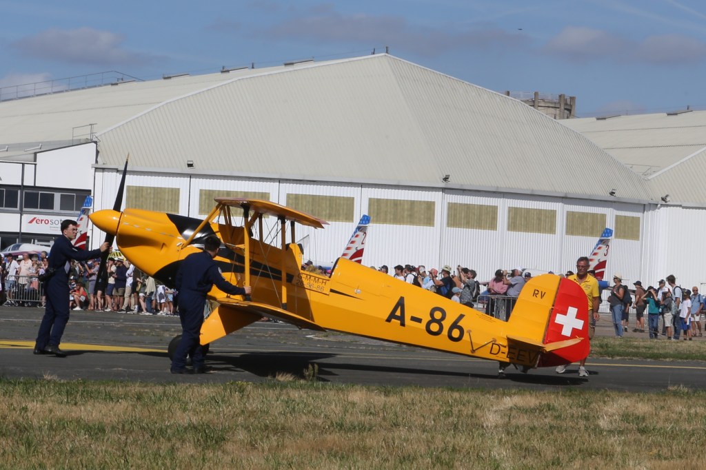 Twee personen in blauwe overalls starten de propeller van een gele vintage biplane met het registratienummer A-86 op een luchtshow. Op de achtergrond is een menigte toeschouwers en een loods te zien.