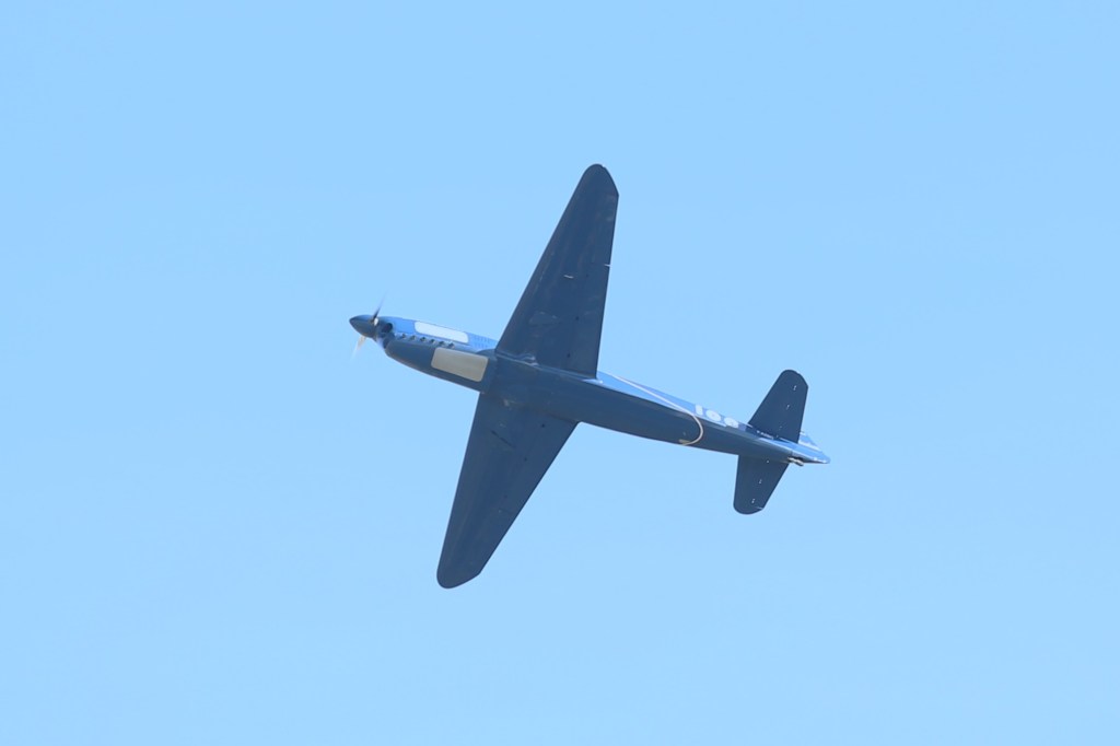Een blauw vliegtuig vliegt hoog in de lucht tegen een heldere, blauwe achtergrond.