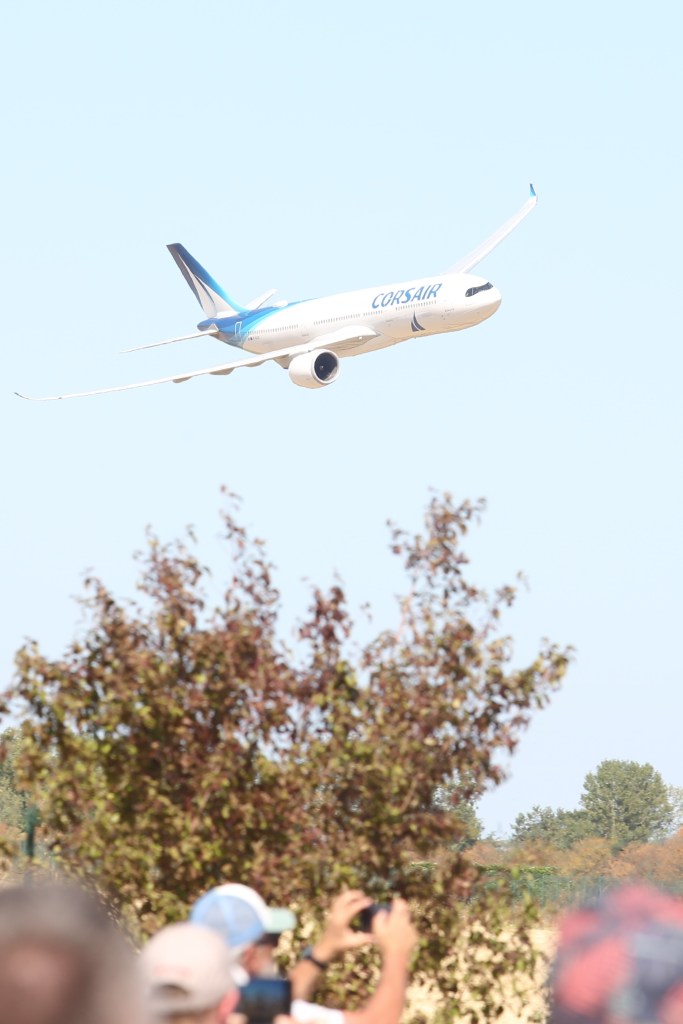 Een Corsair-vliegtuig vliegt hoog boven een menigte, met bomen op de voorgrond.
