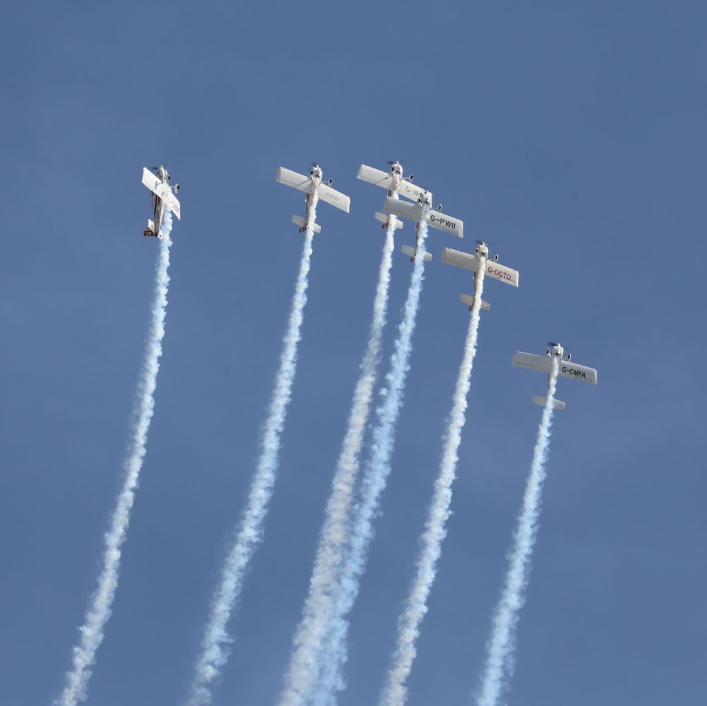 Vijf kleine vliegtuigen die een luchtshow uitvoeren met rooksporen in de lucht.