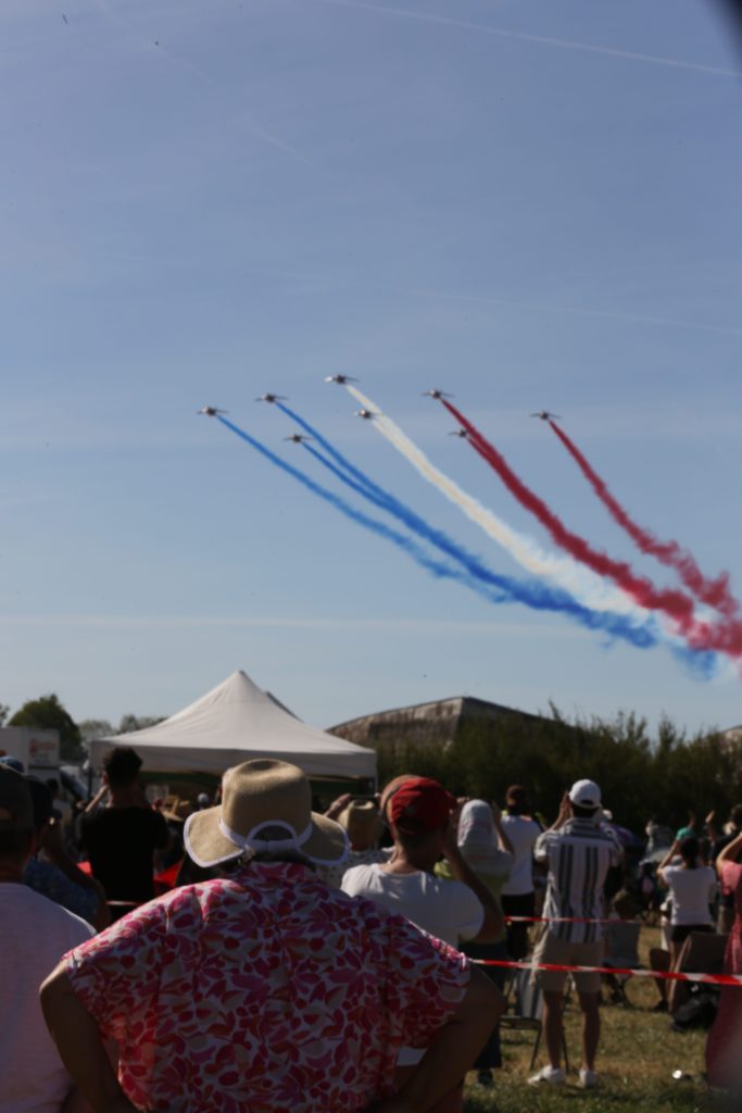 Vliegtuigen in formatie met rook in de kleuren blauw, rood en wit tijdens een luchtshow, met toeschouwers die naar boven kijken.