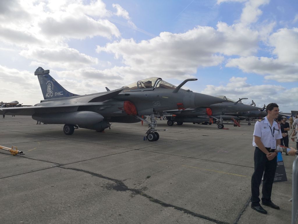Een militaire jet staat geparkeerd op een luchtvaartshow, met andere vliegtuigen op de achtergrond en bezoekers die met personeel praten.