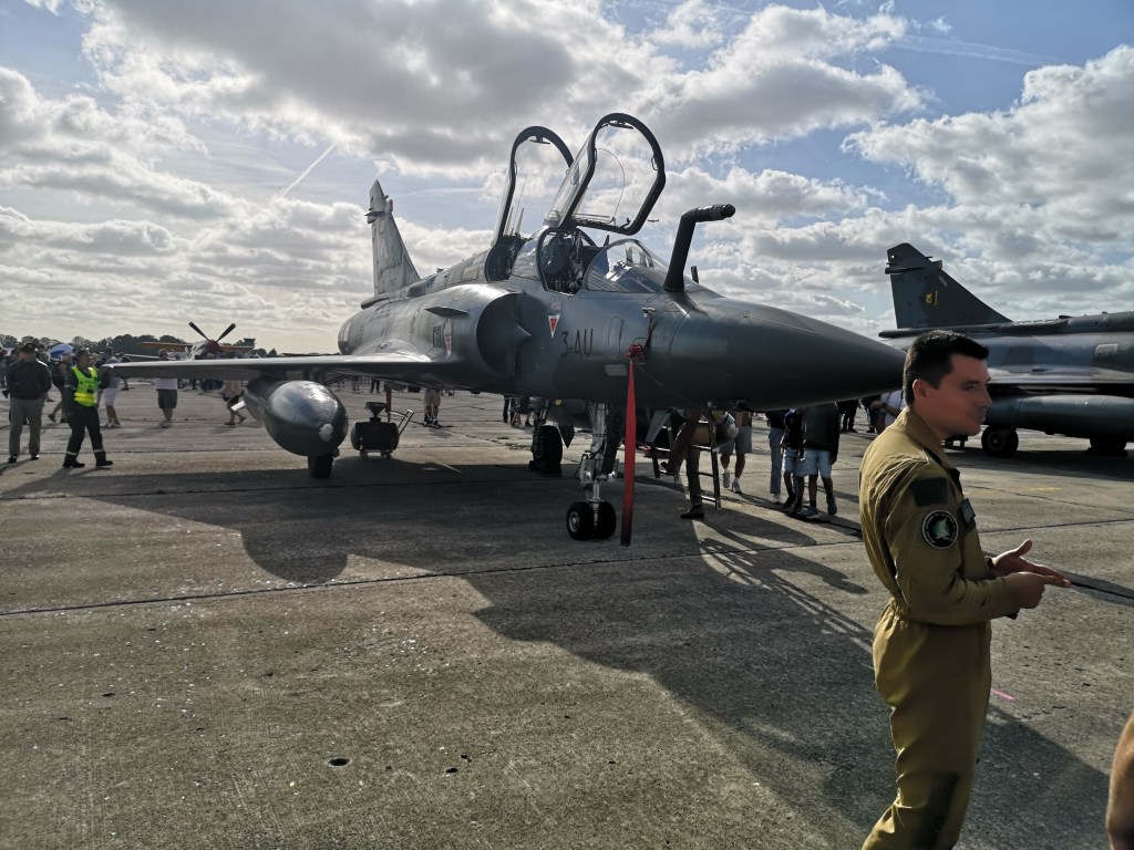 Een jachtvliegtuig met open cockpit staat op een luchtvaartshow, omgeven door bezoekers en personeel in militaire uniformen.