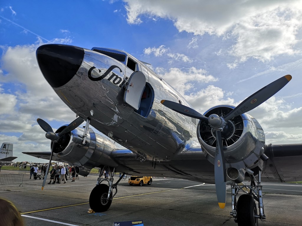 Een vintage vliegtuig met een glanzende metalen romp en propellers, staat geparkeerd onder een heldere lucht met enkele wolken.