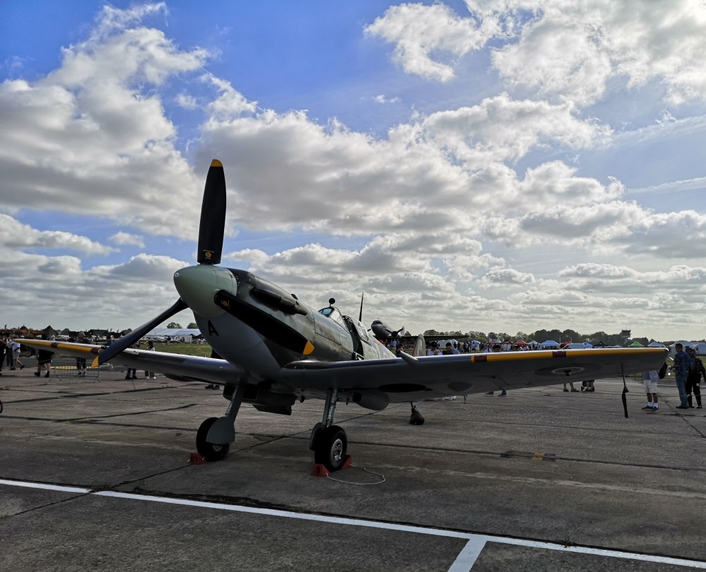 Een historische jager, een Spitfire, staat geparkeerd op een luchtvaartshow met mensen op de achtergrond en een blauwe lucht met wolken.