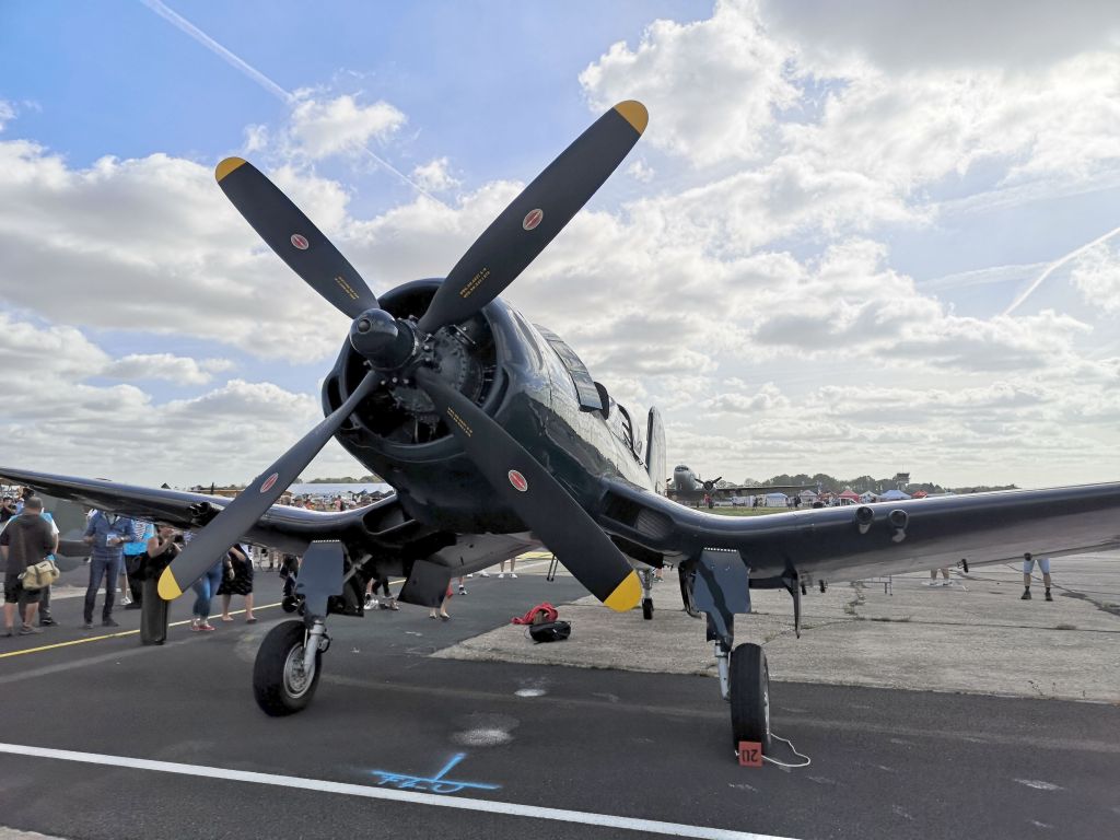 Een vintage vliegtuig met een grote propeller, gefotografeerd op een luchthoogte met mensen op de achtergrond.