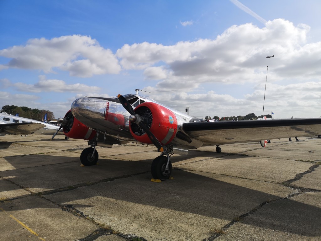 Een vintage vliegtuig met een glanzende metalen romp en rode details staat geparkeerd op een vliegveld onder een heldere lucht met enkele wolken.