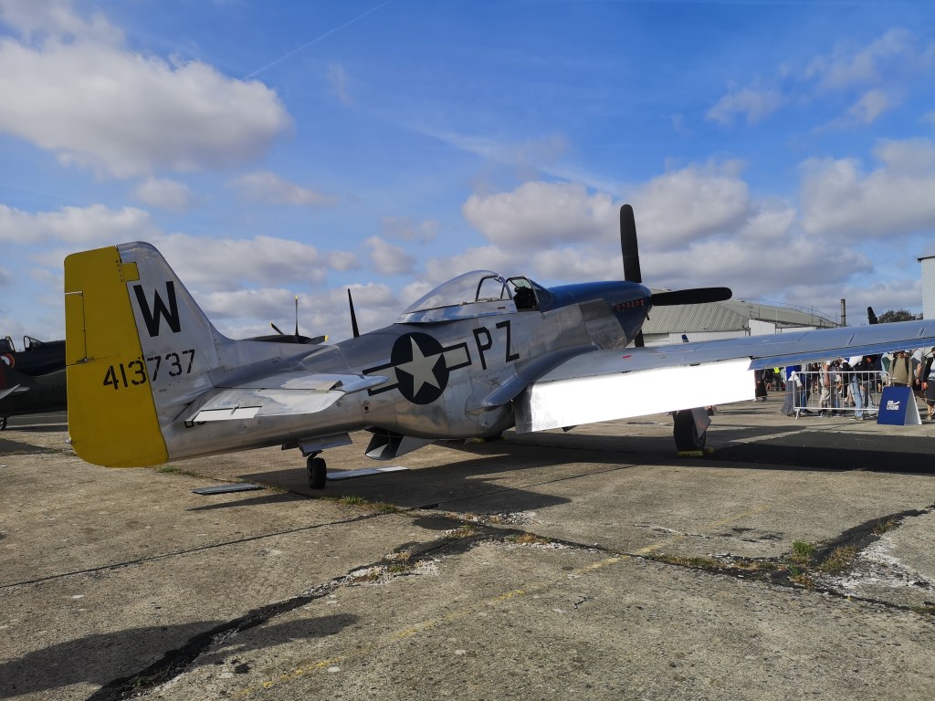 Een historische jachtvliegtuig, de P-51 Mustang, gezien van opzij met het gele staartnummer W en registratie 413737. Op de achtergrond zijn mensen te zien die naar het vliegtuig kijken.