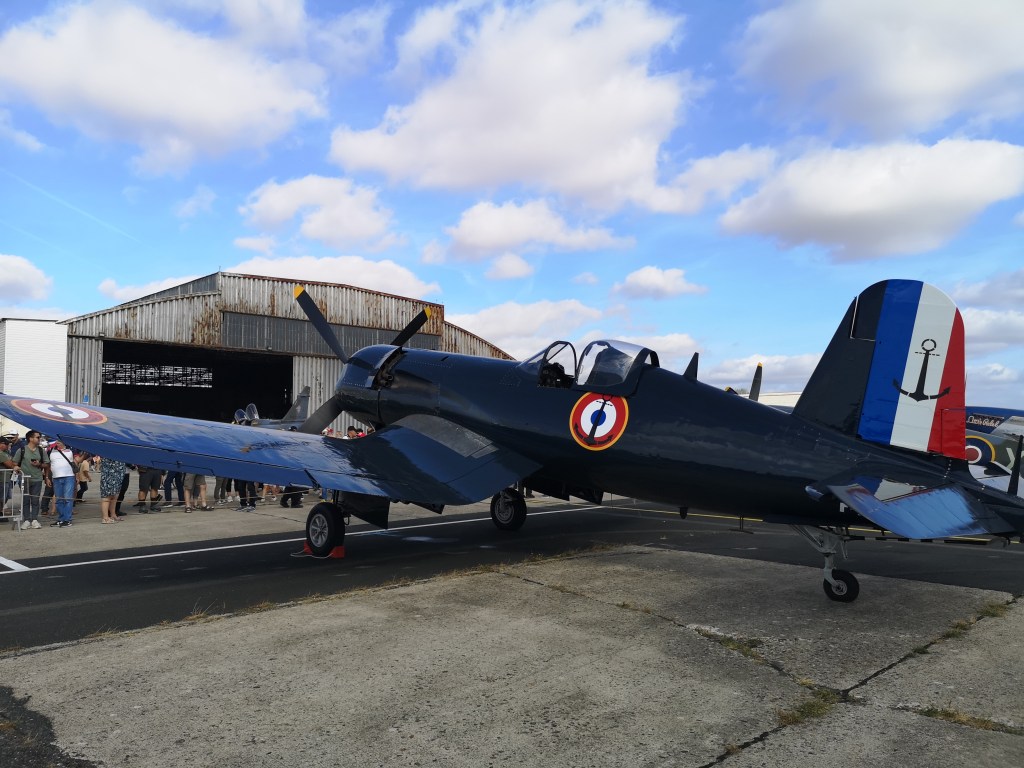 Een vintage vliegtuig met blauwe en witte details staat geparkeerd voor een hangar, omringd door een menigte van mensen.