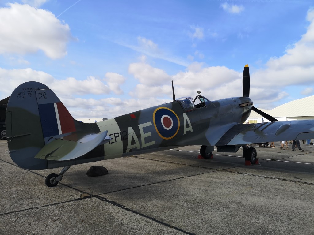 Een historische jachtvliegtuig, een Spitfire, staat geparkeerd op een luchtvaartterrein onder een blauwe lucht met wolken.