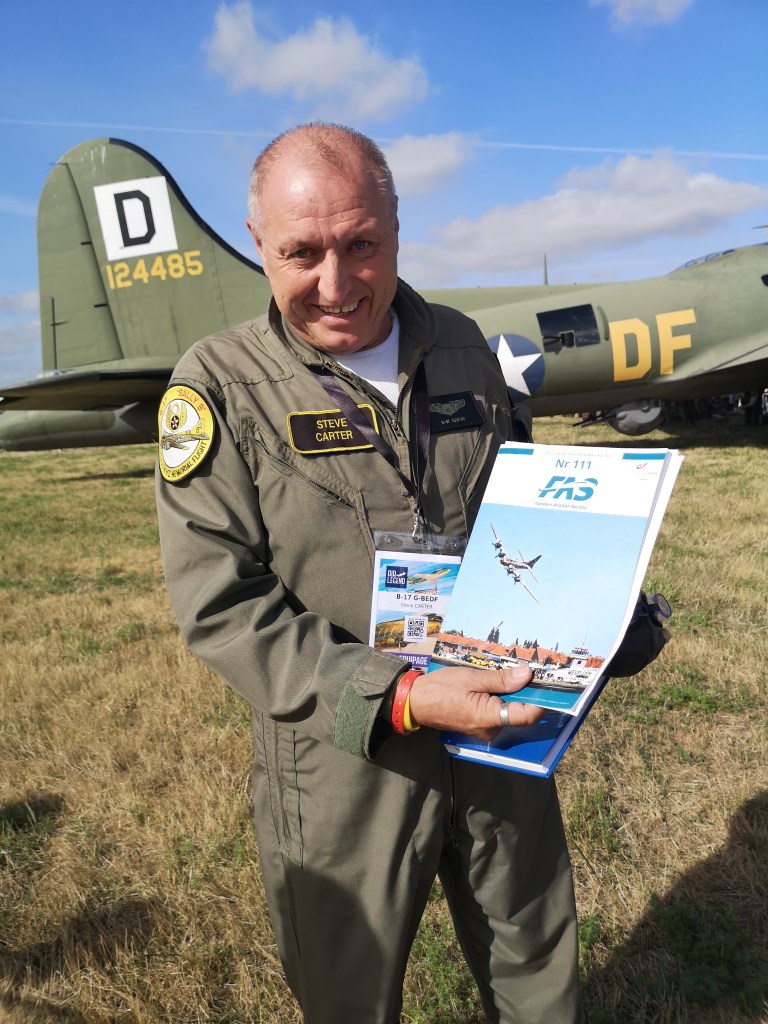 Een man in een vliegersuniform houdt een tijdschrift vast met een afbeelding van een vliegtuig. Achter hem staat een oude militaire bommenwerper met de registratie D124485.