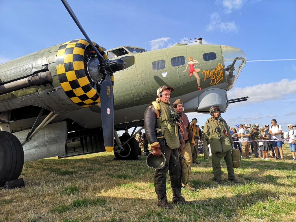 Drie mannen in vintage luchtvaartuniformen staan naast een historische bommenwerper met een uitgesproken neus en een afbeelding van een vrouw op de zijkant, op een evenement met een menigte op de achtergrond.