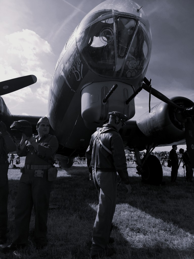 Een groep mensen in vintage pilotenuniformen staat voor een grote oude militaire bommenwerper, met een teddybeer zichtbaar in de cockpit.
