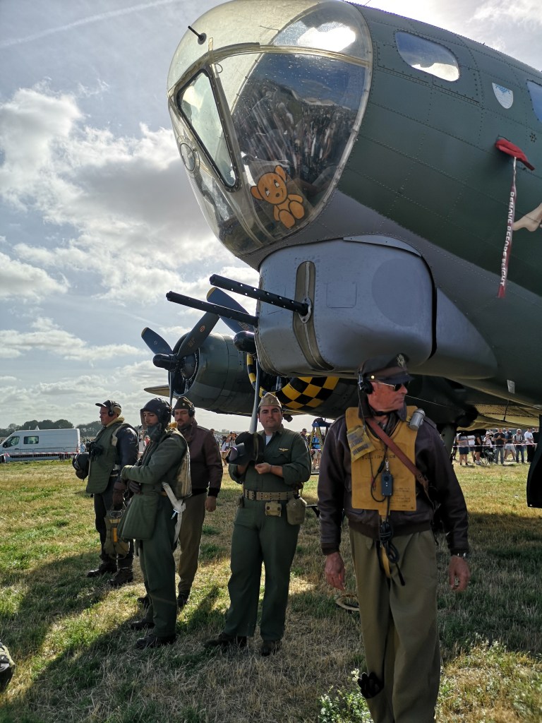 Een historische bommenwerper met een knuffelbeer in de cockpit en bemanningsleden in militaire uniformen die erbij staan.