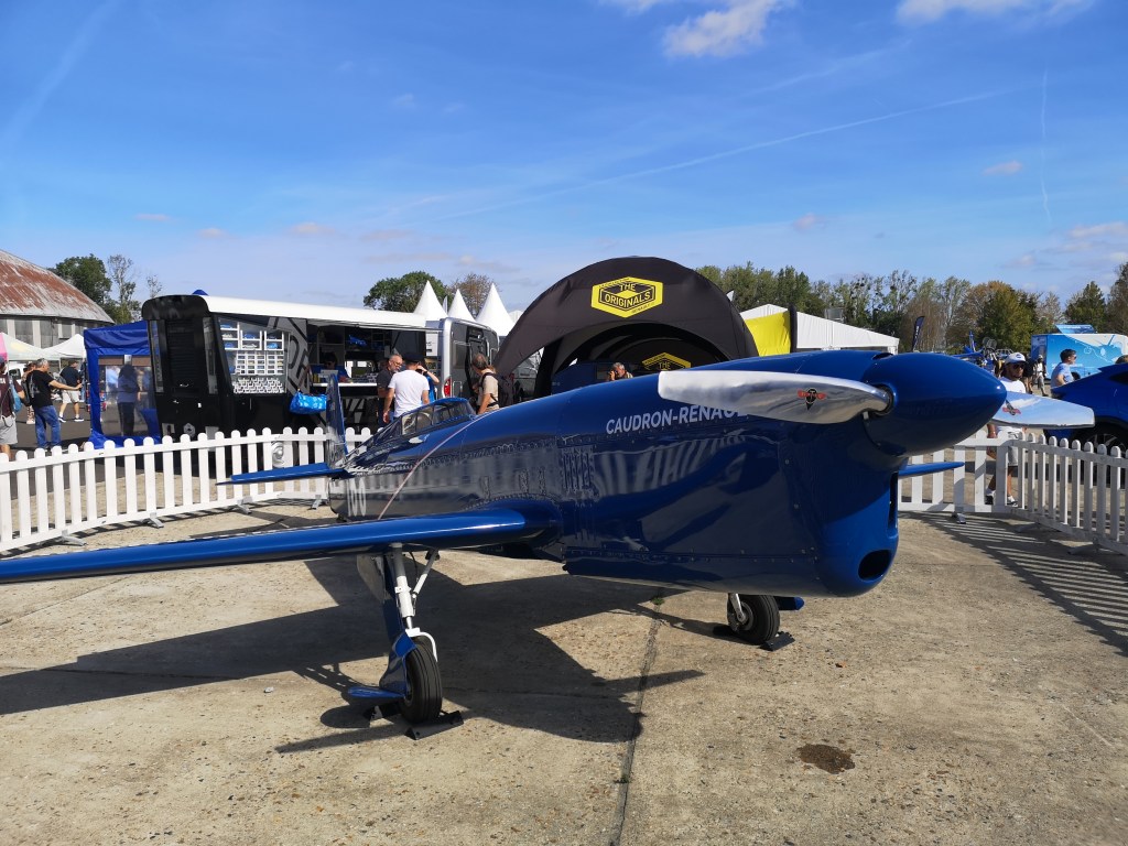 Een blauwe lichte vliegtuig met de naam 'Caudron-Renault', te zien op een luchtvaartbeurs met bezoekers op de achtergrond.