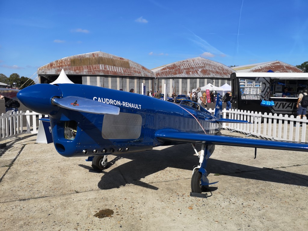Een Caudron-Renault vliegtuig in een luchtshow, met een blauwe kleur en een achtergrond van windtunnels en mensen.