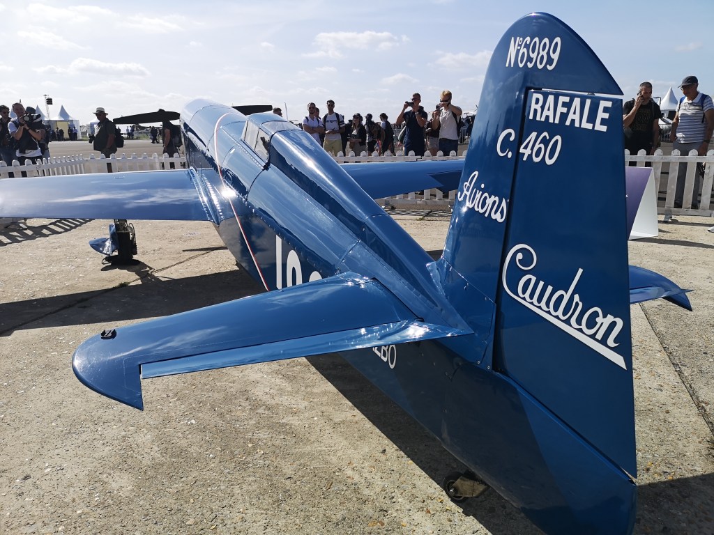 Een blauw Caudron Rafale C.460 vliegtuig met de registratie N°6989, bekeken vanuit de achterkant, met mensen op de achtergrond die het toestel observeren.